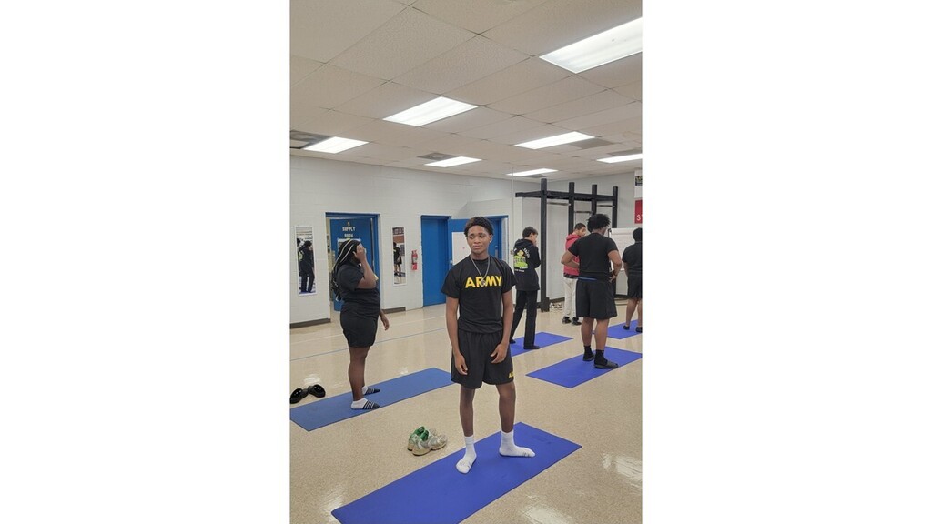 Group of people on yoga mats, standing and seated, in a room with blue walls, white ceiling, and overhead lighting.