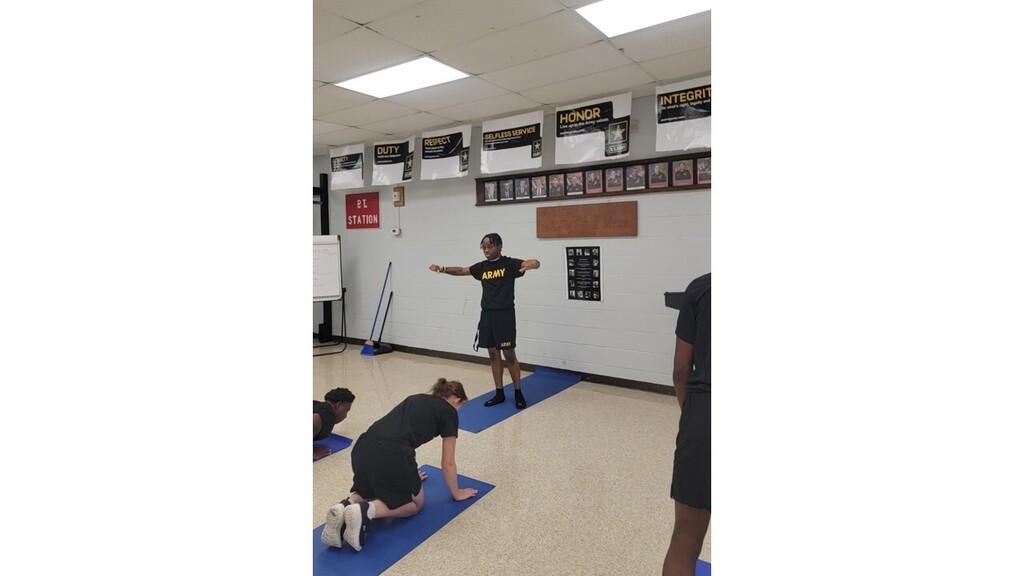 Individuals in a room, one standing with arms extended, another kneeling on a mat, and others nearby.