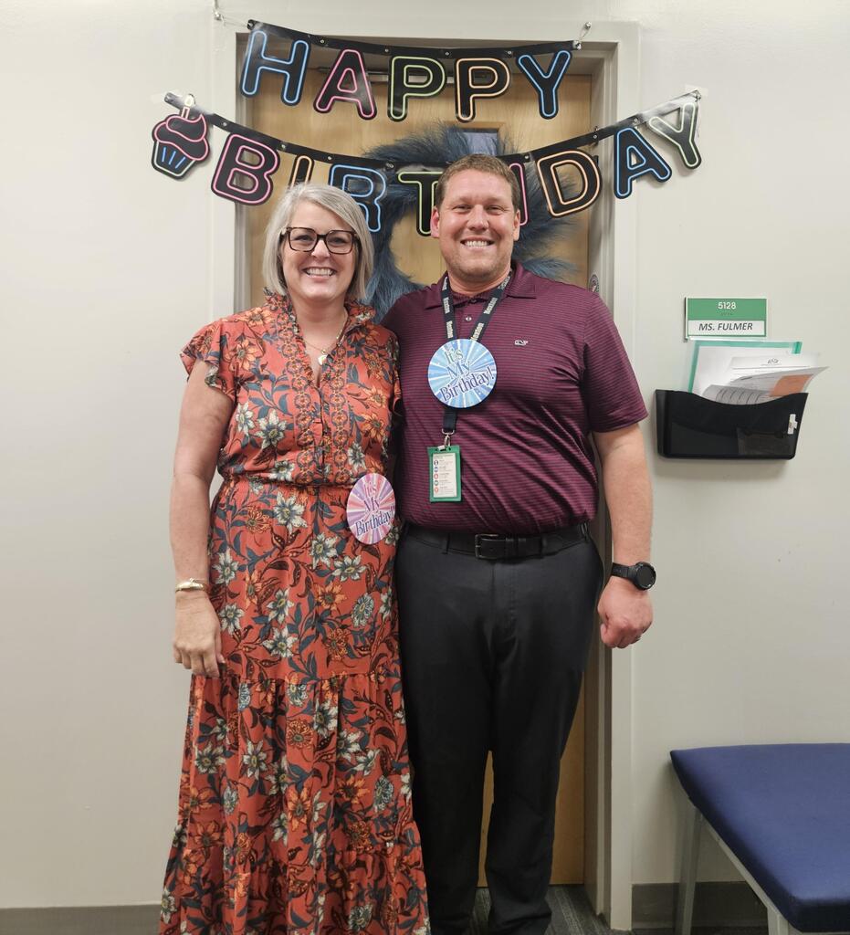 Two people stand next to each other under a banner that says "Happy Birthday." Both wear name tags.