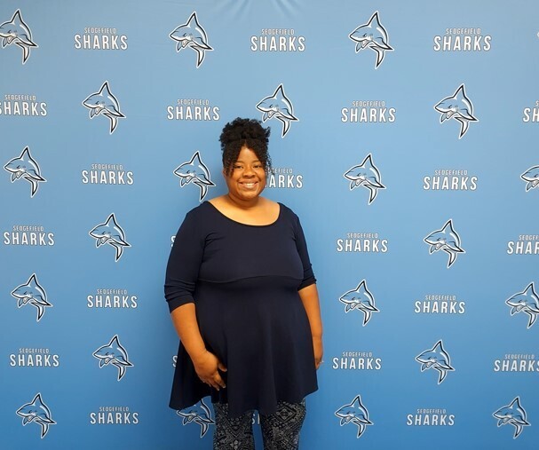 Person in a navy dress standing in front of a blue background with shark images and text.