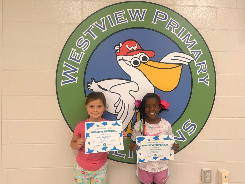 Two young girls holding certificates stand in front of a school logo featuring a bird.