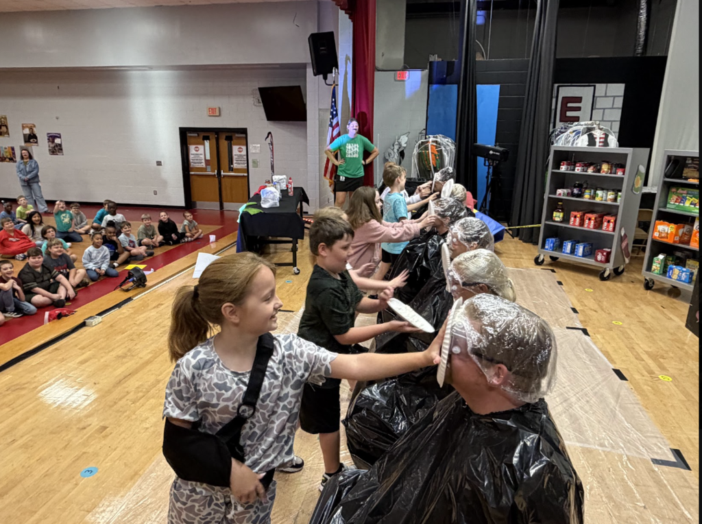 A room with wooden floors where a person gets their hair washed. A group of kids and adults are gathered around.