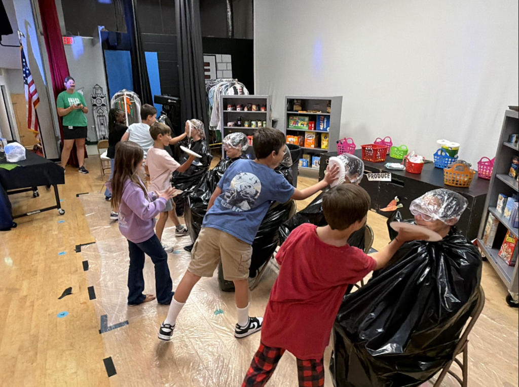 Kids with hair covered in foil participate in a school event. Behind them, a table holds plastic bags and a bookshelf.