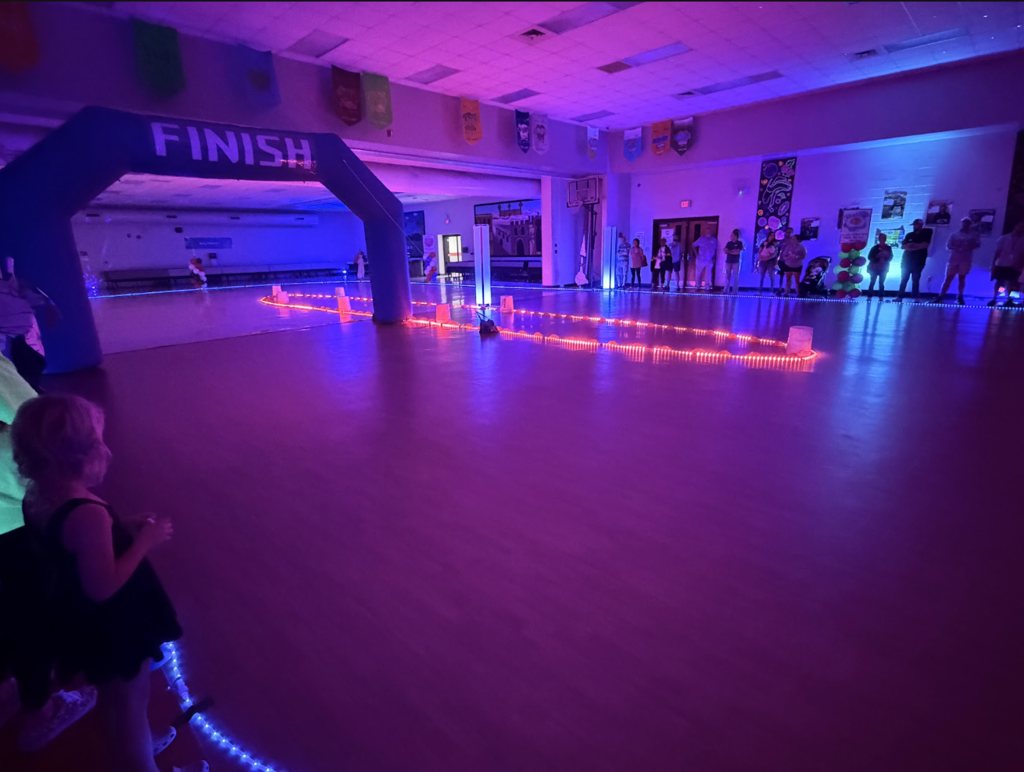 An indoor track for a children's race with a finish line, colorful lights, and people watching.