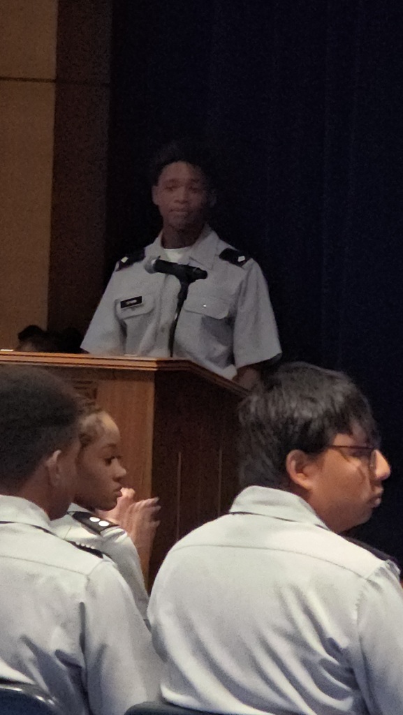 An adult in uniform speaks at a podium; students in uniform listen. Brown wooden podium, black curtain background.