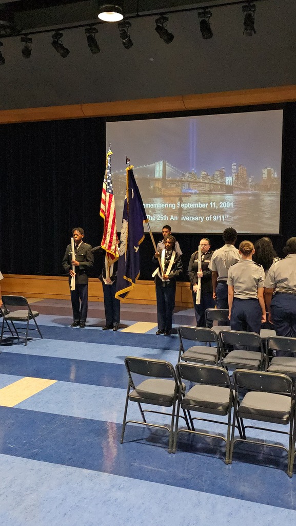 A group of people stand on a stage holding flags. Behind them is a large screen displaying an image. Chairs are arranged in front of them.
