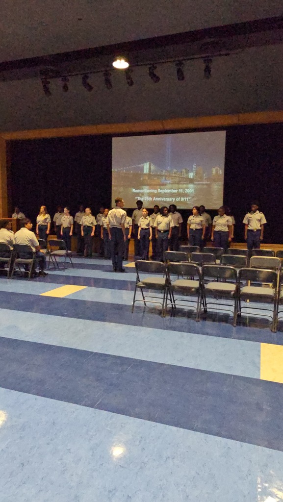 A group of people stand on a stage in front of a projector screen. Chairs are arranged in rows.