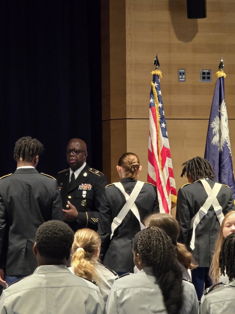 Several individuals in gray uniforms stand before a stage with American and state flags. A man in military uniform addresses the group.