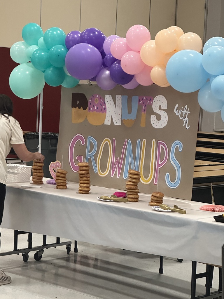 A table with a white tablecloth displays doughnuts and a sign reading "Donuts with Grownups". Colorful balloons float above.