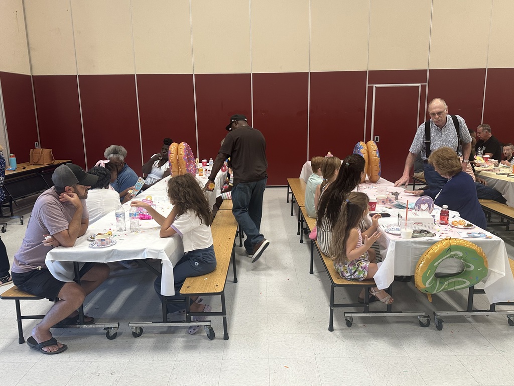 People seated at tables with food. Some are adults and some are children. The room has white and red walls.