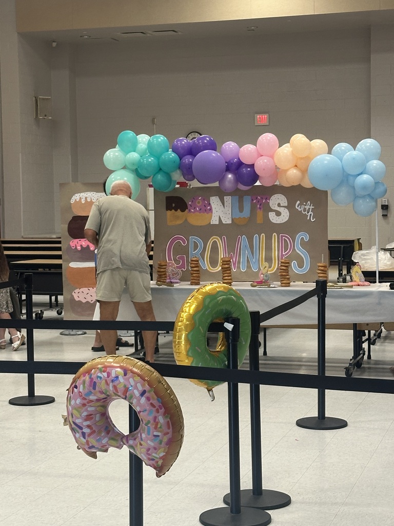 A man stands behind a table with a colorful balloon arch and signs reading "Donut Groups." Two oversized donuts are in front.