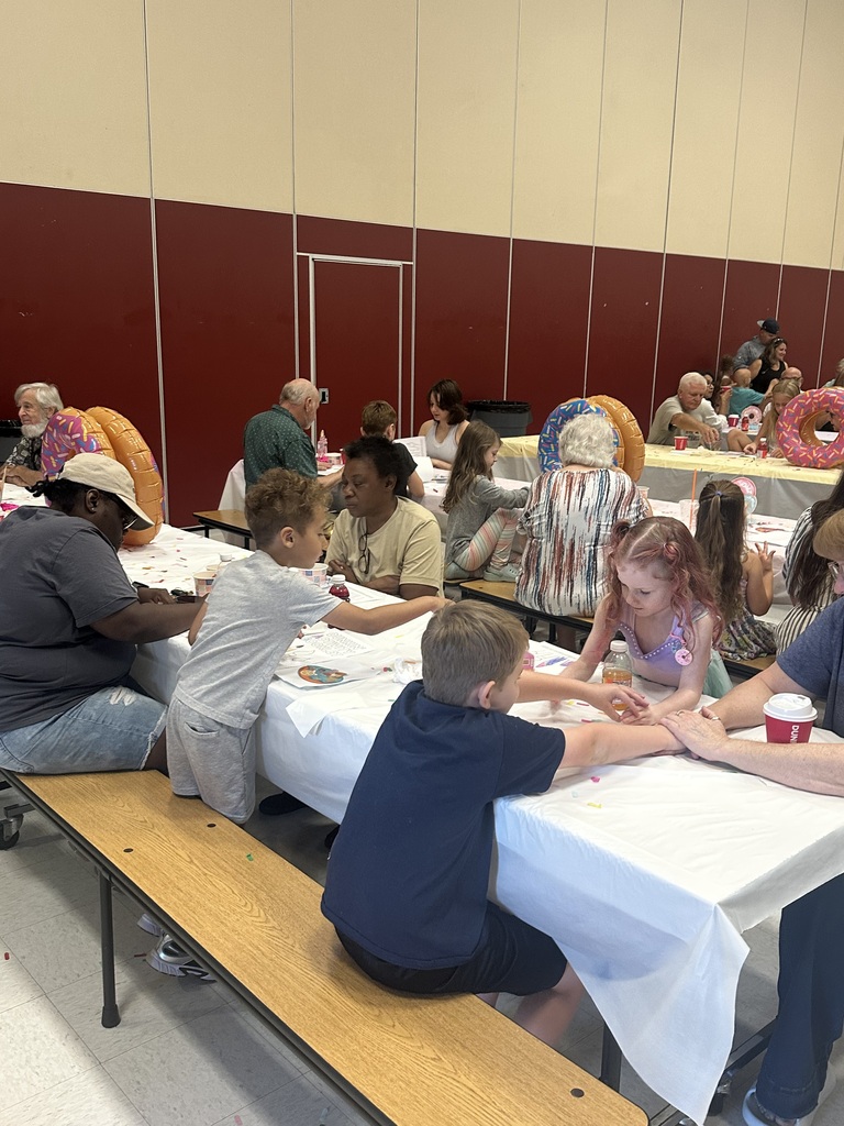 A group of people sit at long tables in a room, engaged in an activity. The tables have white tablecloths.