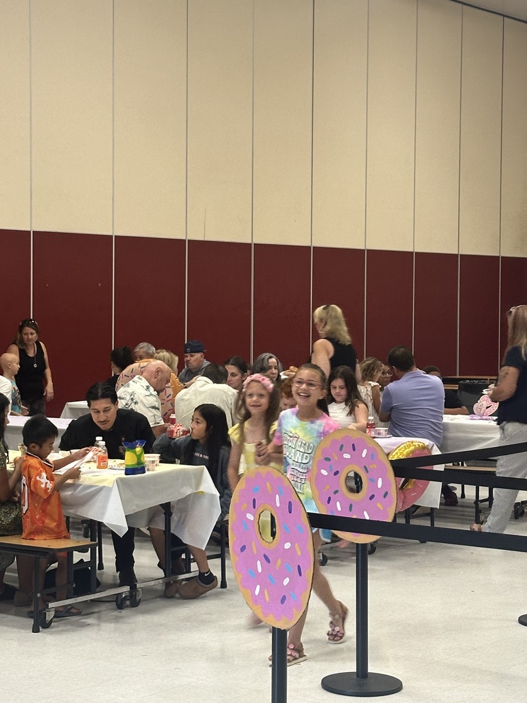 A group of people gathered around tables in a room, with one child walking between tables. Some are seated, while others are standing.