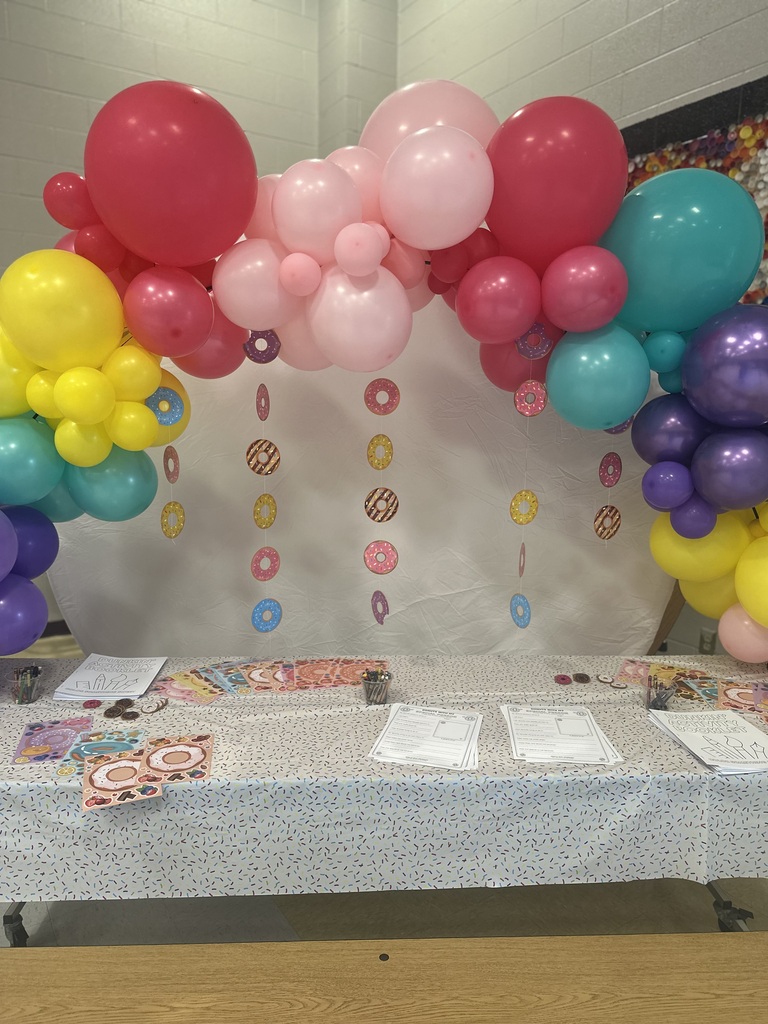 A table with a white tablecloth holds papers and balloons. Balloons in various colors float above, and decorative items hang from the ceiling.