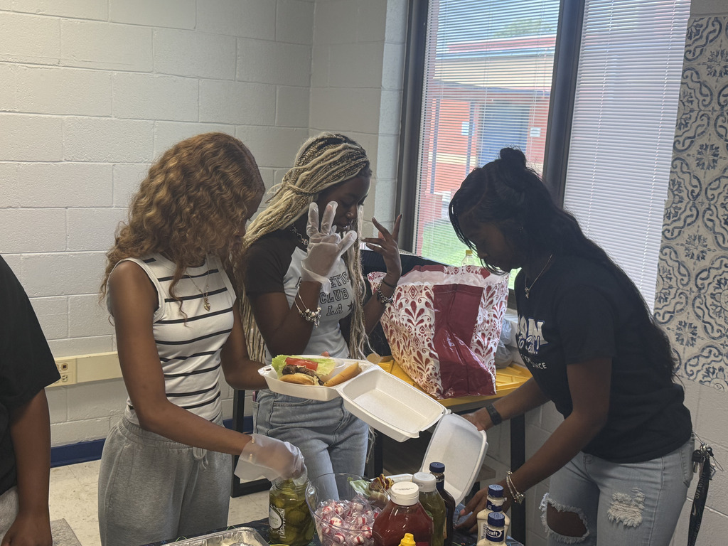Three people in a classroom prepare food. A table is laden with bottles of condiments and a sandwich.