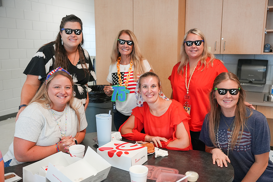 Group of six women wearing sunglasses and smiling, sitting around a table with food and drinks.