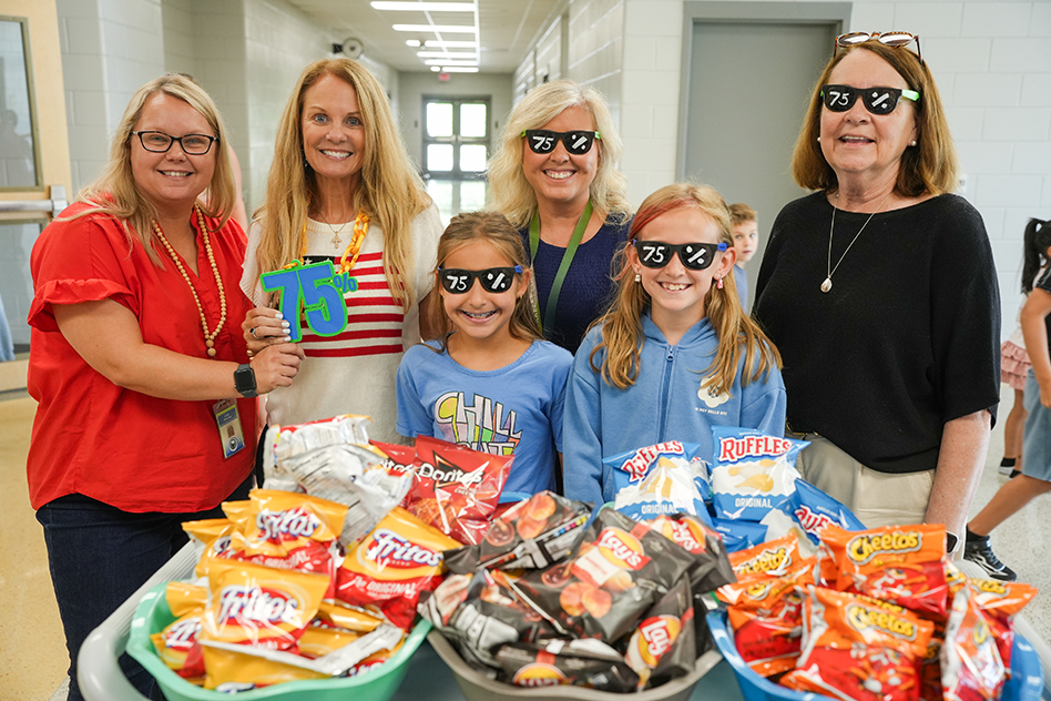 Adults and two girls, one wearing sunglasses, stand behind a table with bags of snacks.