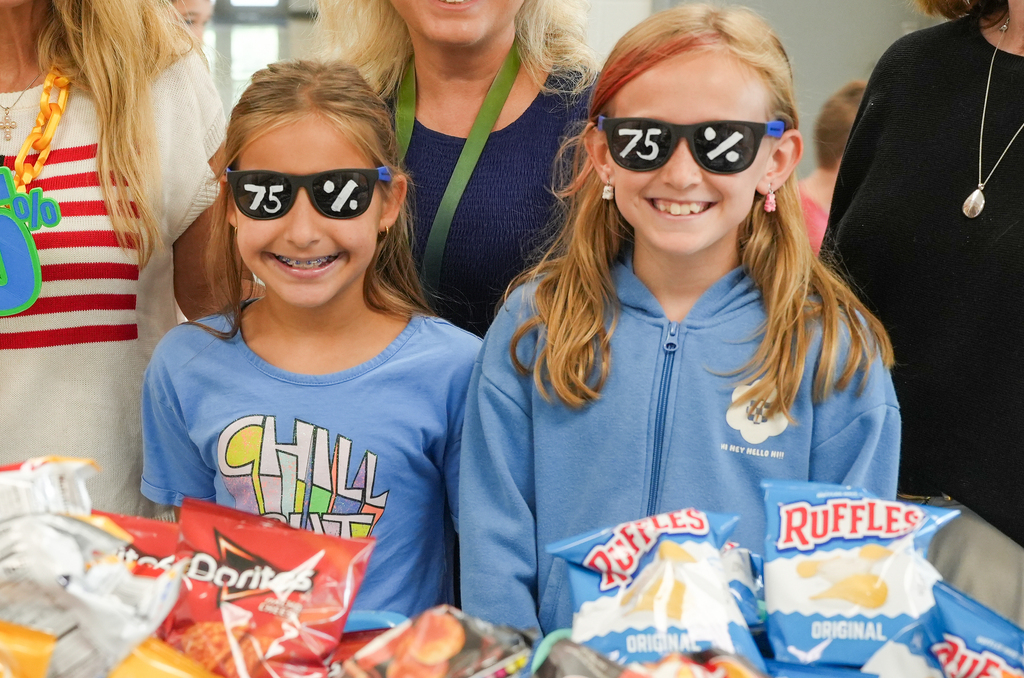Two girls wear sunglasses and smile. They stand by a table of snacks. People behind them.