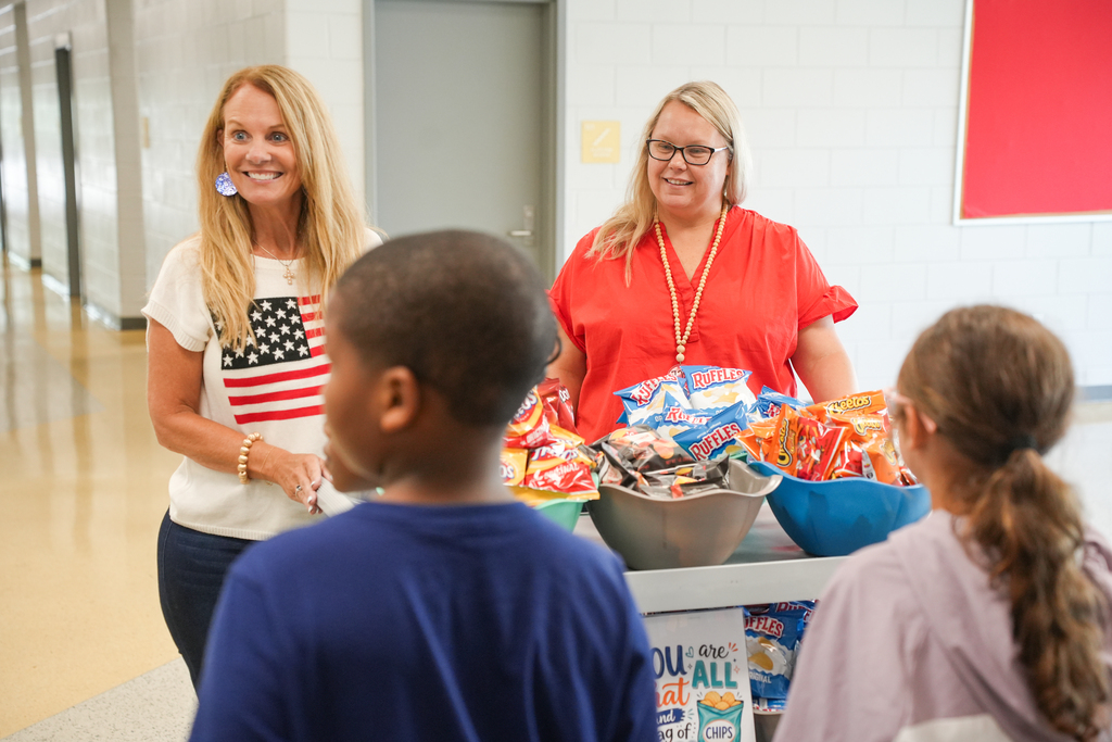 Two women and two children gather around a table with snacks. One woman wears glasses and a lanyard, another an American flag shirt.