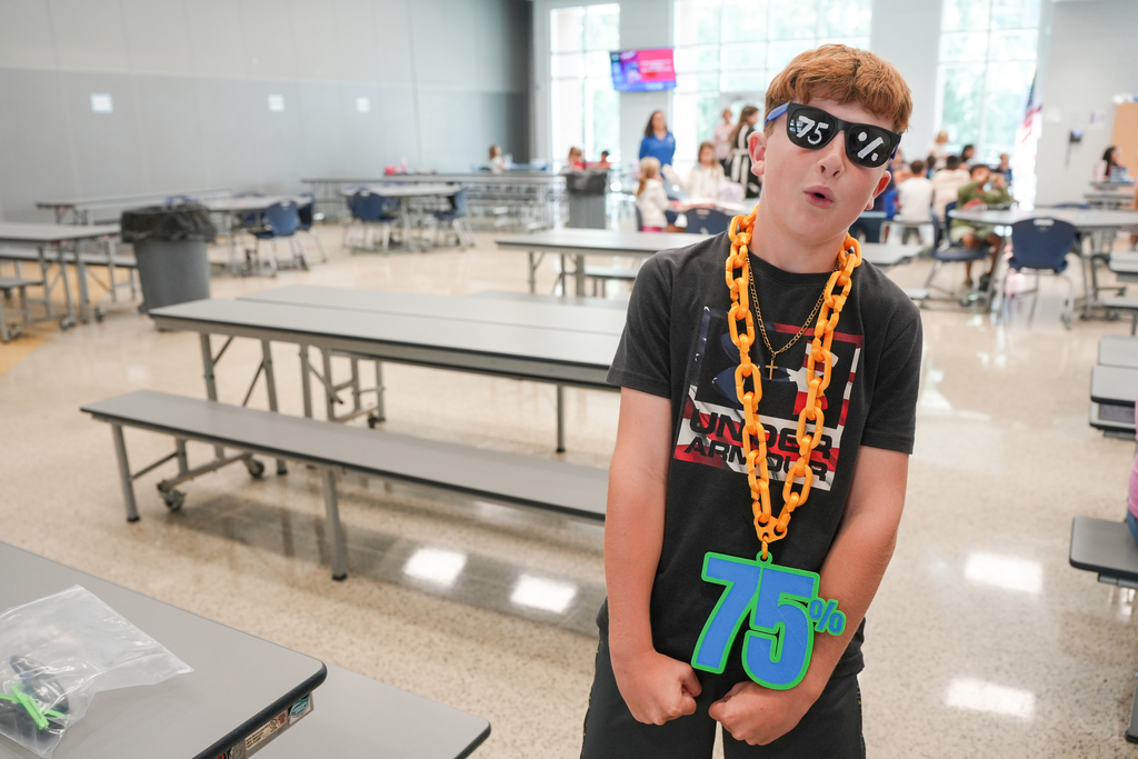 Boy wearing a necklace with a large number 75, sunglasses, and a black shirt stands in a school cafeteria.