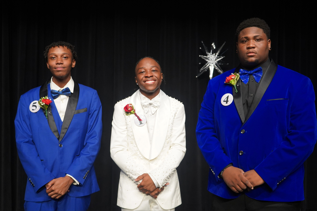 Three men in tuxedos with flowers stand on a stage with a dark background.