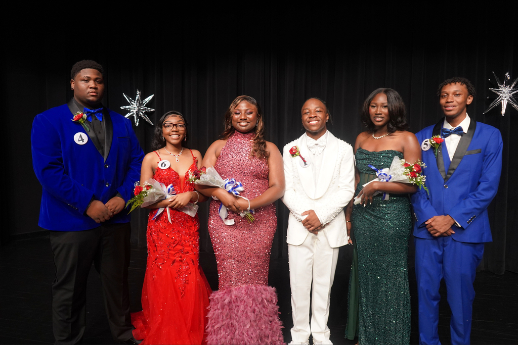 Five people in formal attire stand on stage, holding flowers. The background is a dark curtain.