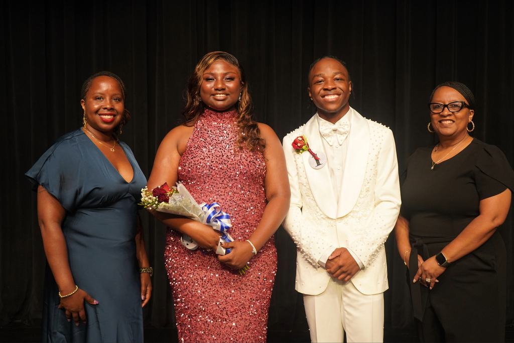 Four adults, one holding a bouquet, stand together against a black backdrop. Two are dressed in formal wear.