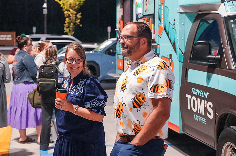 A man and woman, both wearing glasses, stand outside. The man has a floral shirt. A coffee truck is behind them.