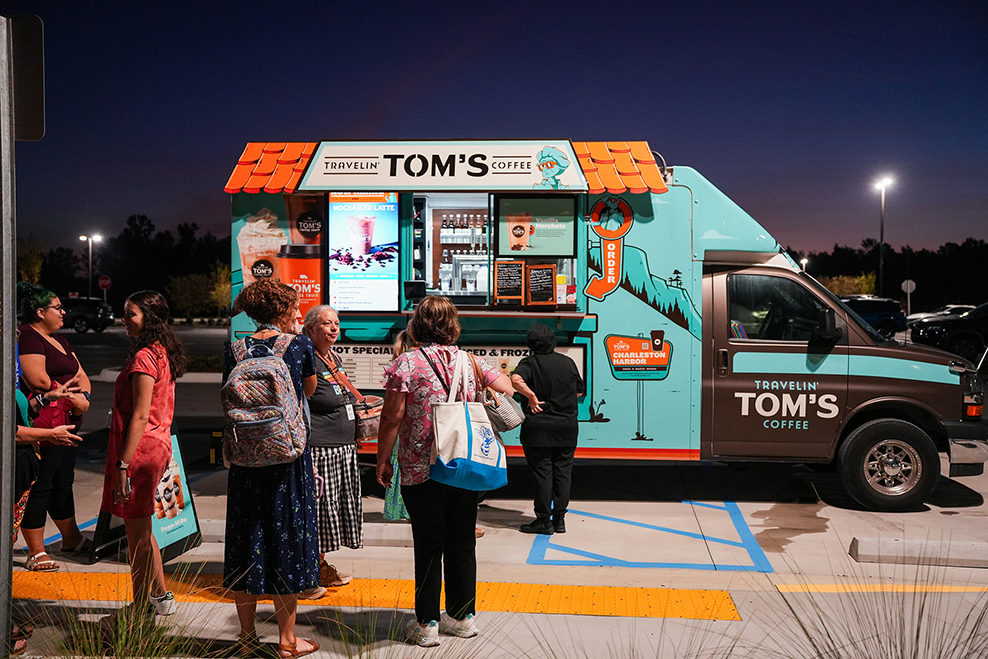 People stand in line in front of a Tom's Coffee food truck, illuminated against a night sky.