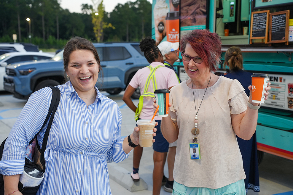 Two women hold drinks and laugh outside a colorful food truck. Other people walk in the background.