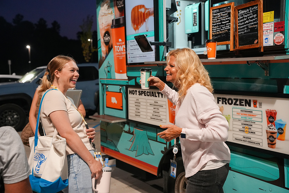 Two women stand before a food truck at night, with one holding a cup and smiling. Behind them, a car is parked.