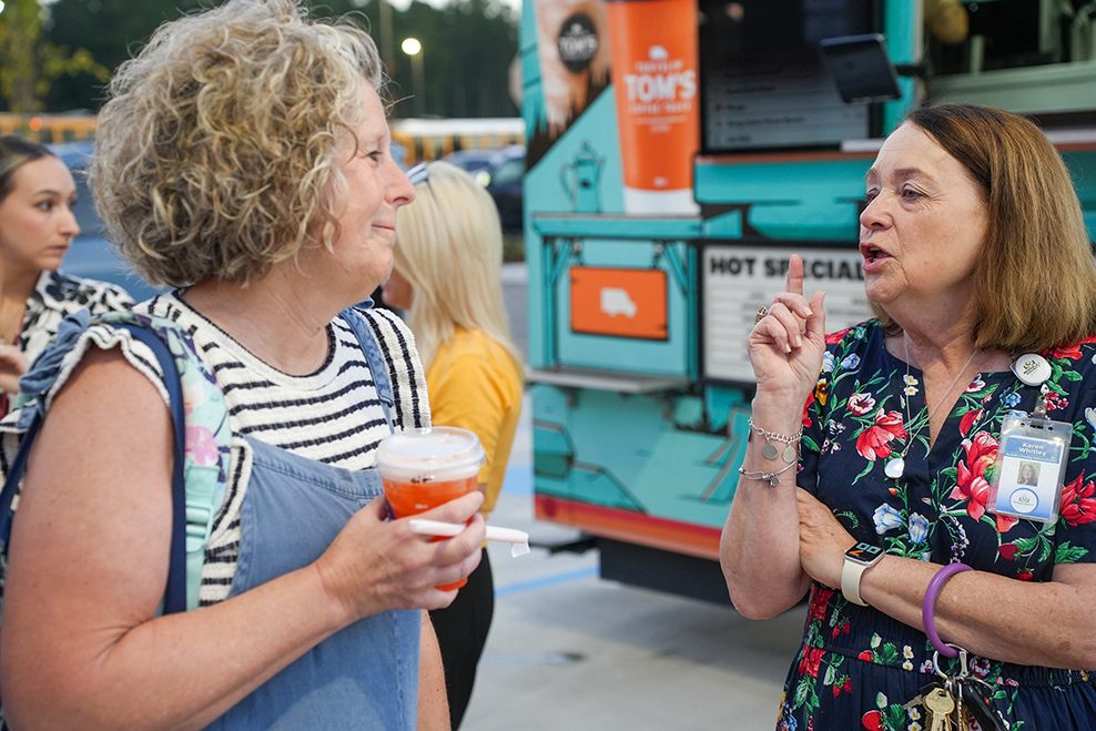 A woman with curly hair and a floral dress holds a drink. Another woman with a backpack speaks to her.
