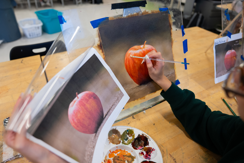 Person paints a pumpkin on canvas using a photo for reference. A paint palette holds colors.