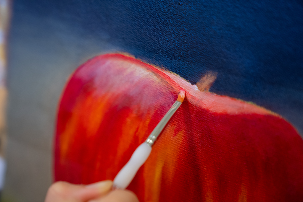 A hand paints a red apple on a blue background. A paintbrush is visible, adding strokes to the apple.
