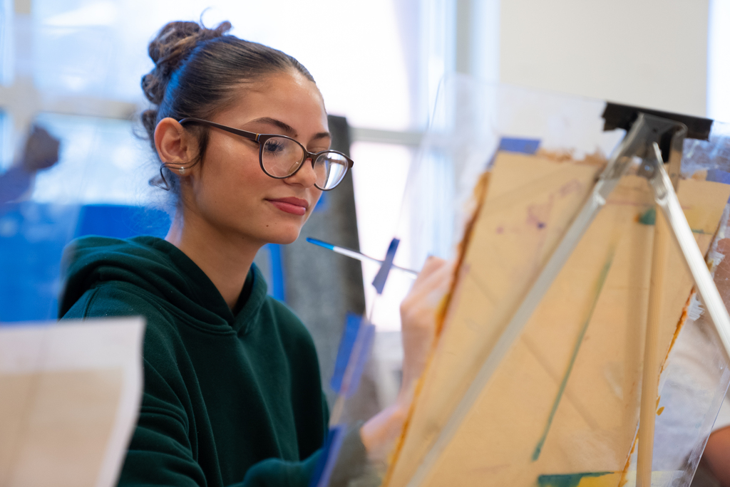 Person in green hoodie with glasses, painting on a slanted cardboard with a brush.