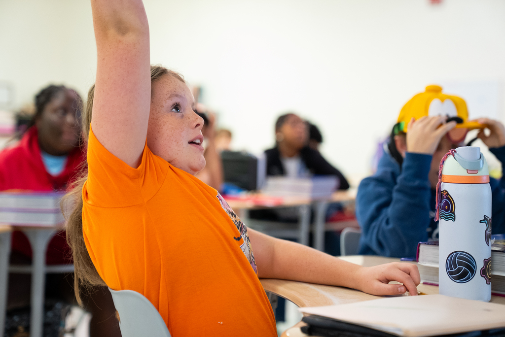 A student in an orange shirt raises their hand in a classroom. Behind, others are seated at desks.