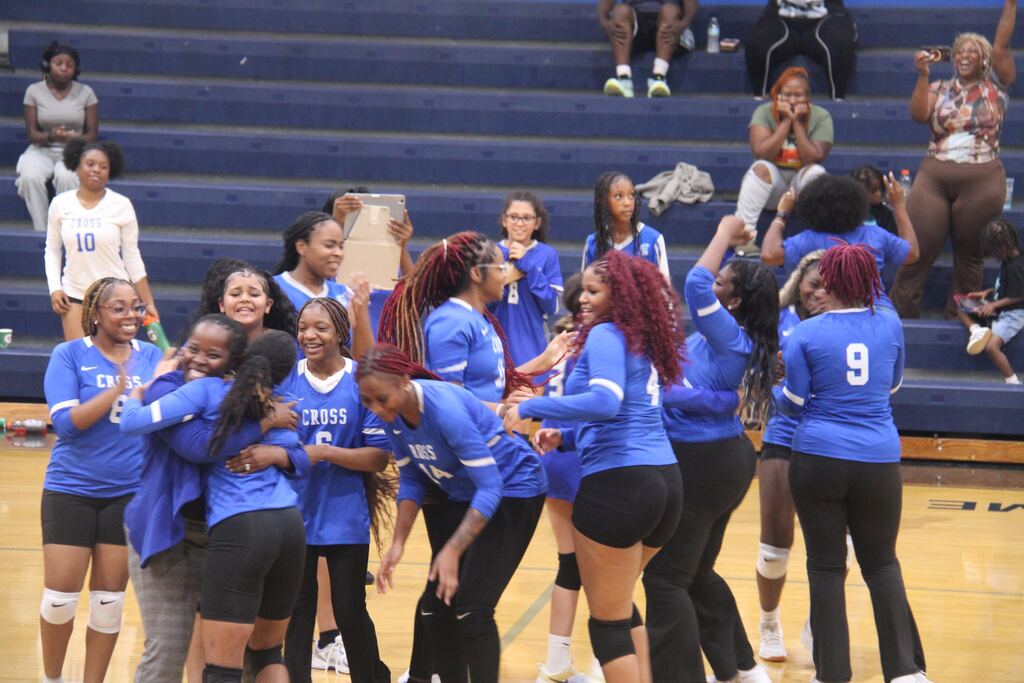 Group of young women in blue uniforms and black shorts huddled together on a court. Audience on bleachers.
