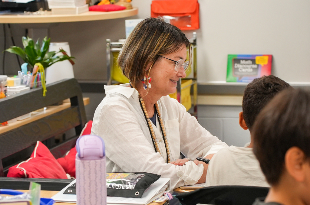 A woman with short hair wearing glasses sits at a desk in a classroom, interacting with a young boy.