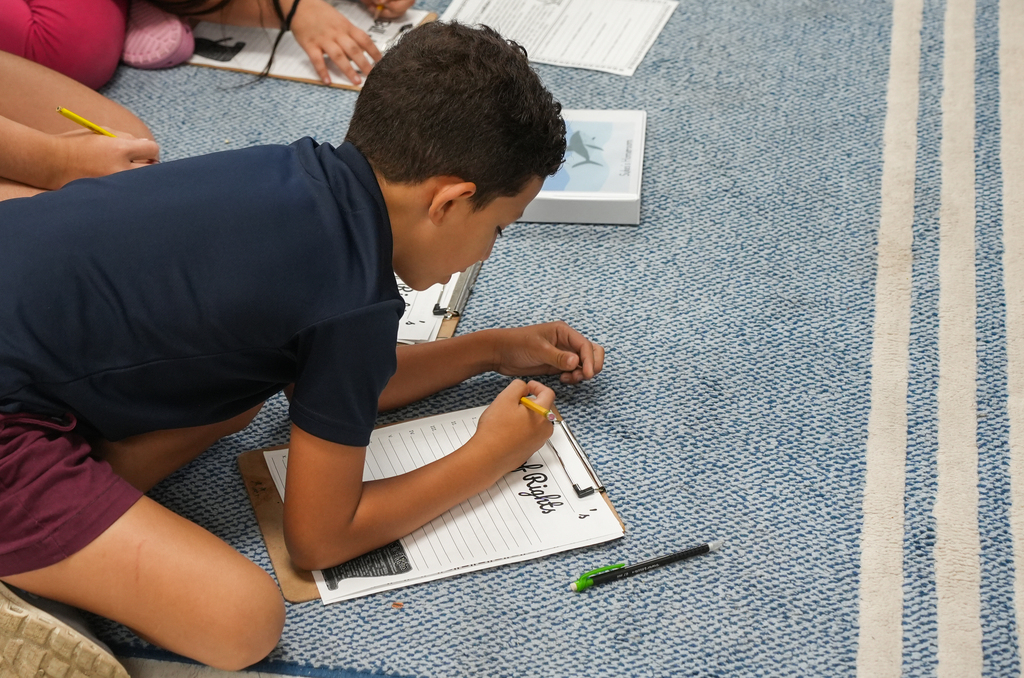 A boy crouches on the floor, writing on a paper. Other children work at their desks in the background.