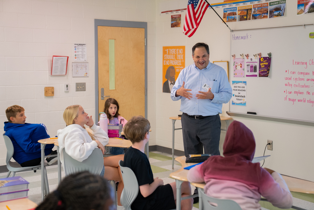 A teacher stands in front of a classroom with students seated at desks. A whiteboard is on the wall.