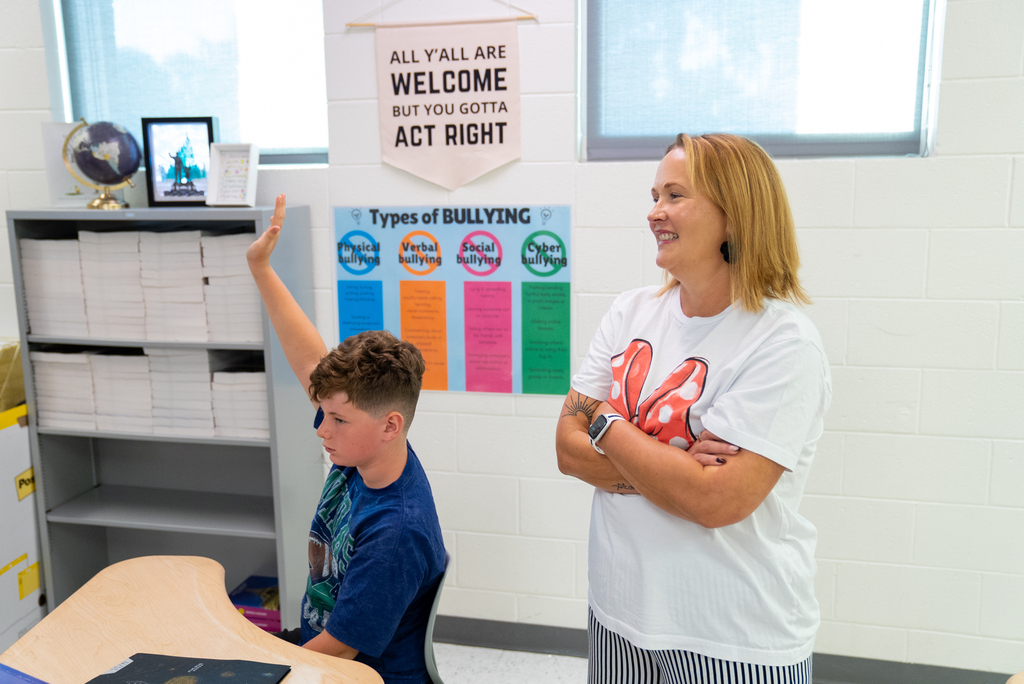 A teacher and student stand in a classroom. The teacher has her arms folded, and the student raises his hand.