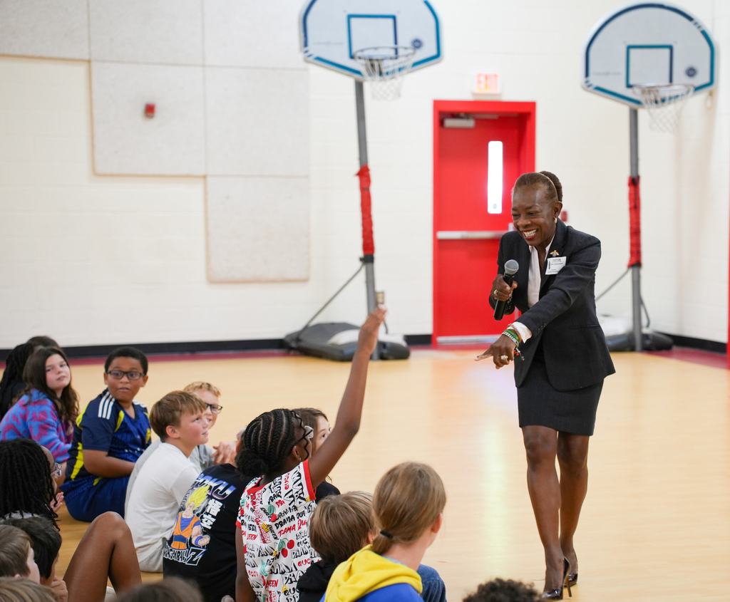Speaker in black suit and skirt standing in gym, holding a microphone, addressing a group of seated children.