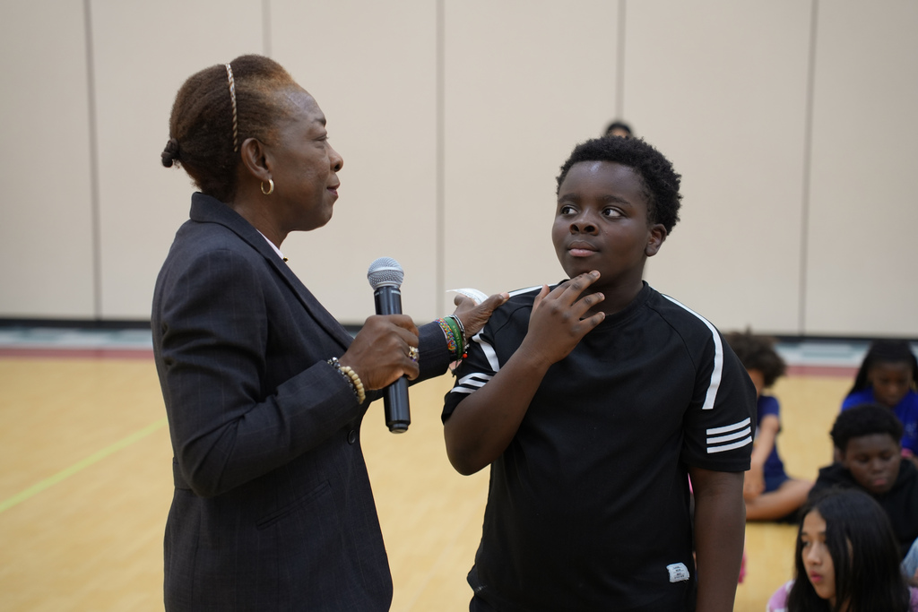 A woman in a black blazer holds a microphone while speaking to a young boy in a black T-shirt in a gym.