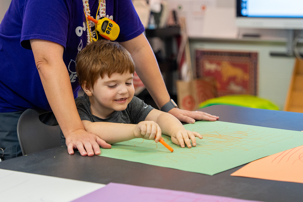 A young boy smiles, drawing on a green paper sheet while a woman helps. Background includes a desk, colorful papers, and a TV monitor.