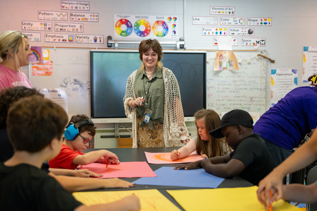 A teacher stands in front of a classroom, guiding students seated around a table with art projects.
