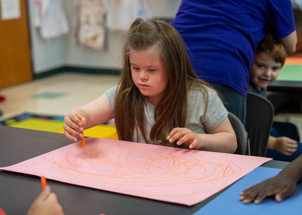 A girl with long hair draws on a pink sheet of paper in a classroom. Another child watches.
