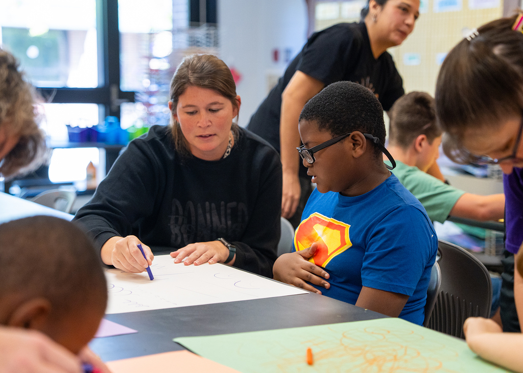 A teacher helps a student with glasses draw on a large paper at a table. Other children and adults are around.