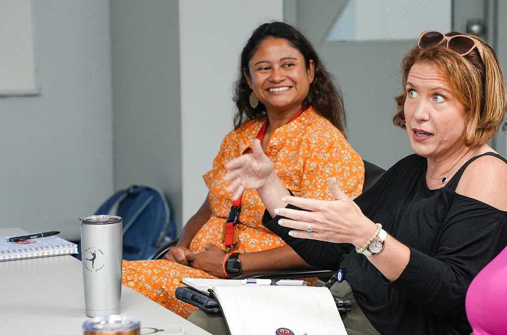 Two women sitting at a table. One smiles and the other talks with hand gestures. A cup, papers, and a backpack are on the table.