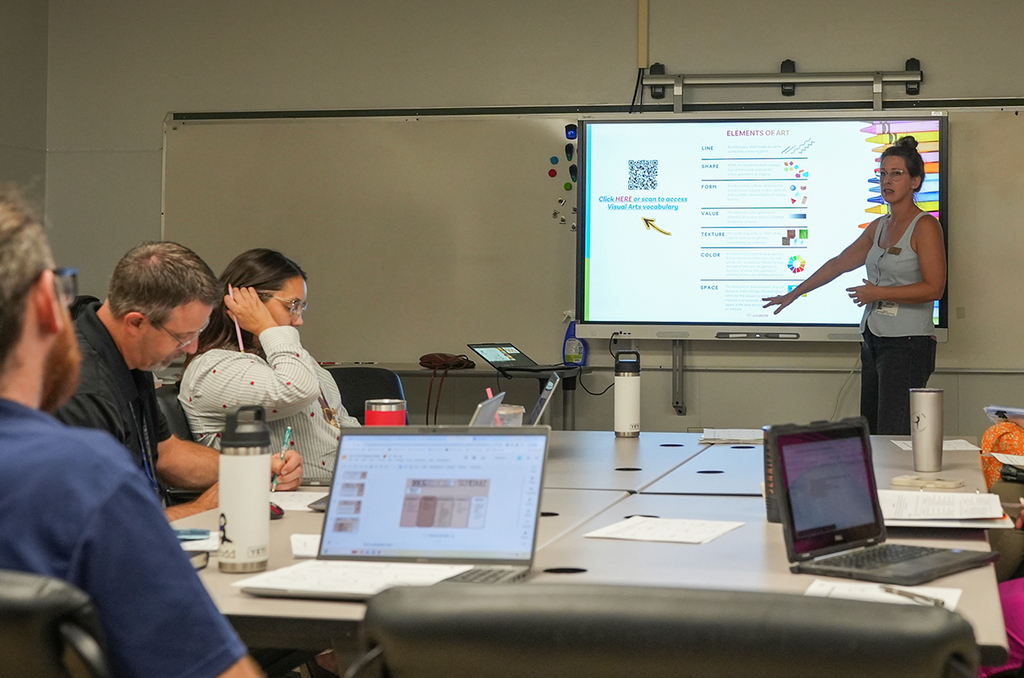 A woman is standing at a table in front of a screen and a whiteboard. Attendees sit around the table with laptops.