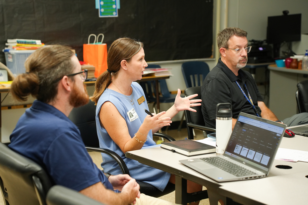 People sit around a desk in a classroom with a laptop, one woman gesturing while speaking.